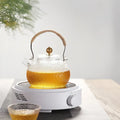 Clear textured glass teapot with woven handle and golden lid knob on white ceramic electric earthenware oven base