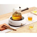Clear glass teapot with dark wooden handle and black lid knob filled with golden tea beside ceramic electric earthenware oven