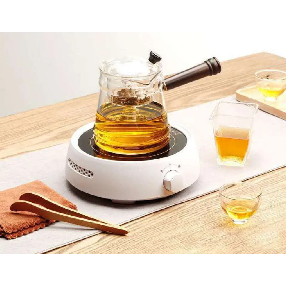 Clear glass teapot with dark wooden handle and black lid knob filled with golden tea beside ceramic electric earthenware oven