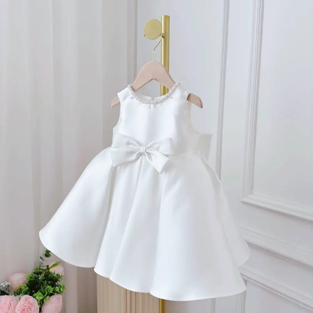 White satin sleeveless baby dress with beaded neckline and large bow in elegant white classic style